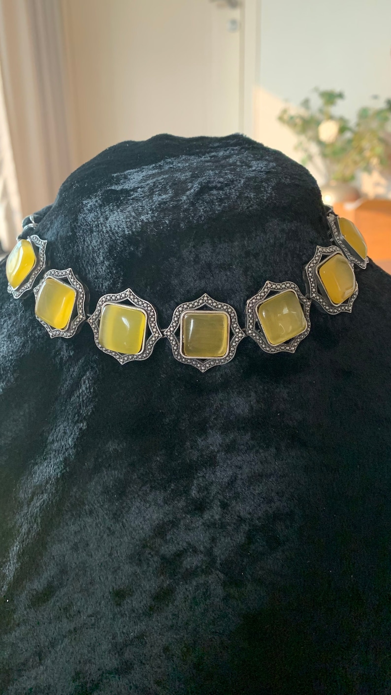 May include: A silver choker necklace with a geometric design and yellow square-shaped stones.