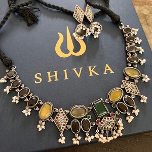 May include: A silver-toned necklace and earring set with oval and diamond-shaped gemstones in shades of yellow, green, and brown. The necklace has a black cord and the earrings have small pearl accents. The jewelry is displayed on a navy blue box with the word "SHIVKA".