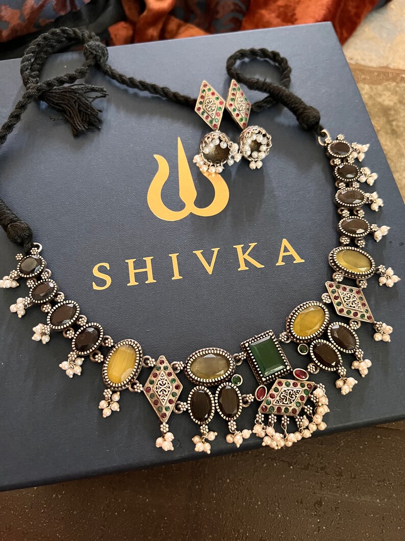May include: A silver-toned necklace and earring set with oval and diamond-shaped gemstones in shades of yellow, green, and brown. The necklace has a black cord and the earrings have small pearl accents. The jewelry is displayed on a navy blue box with the word "SHIVKA".