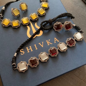 May include: Two silver necklaces with colorful gemstone accents. The first necklace has yellow gemstones and the second necklace has red and white gemstones. Both necklaces are on a blue box with the word "SHIVKA" printed on it.