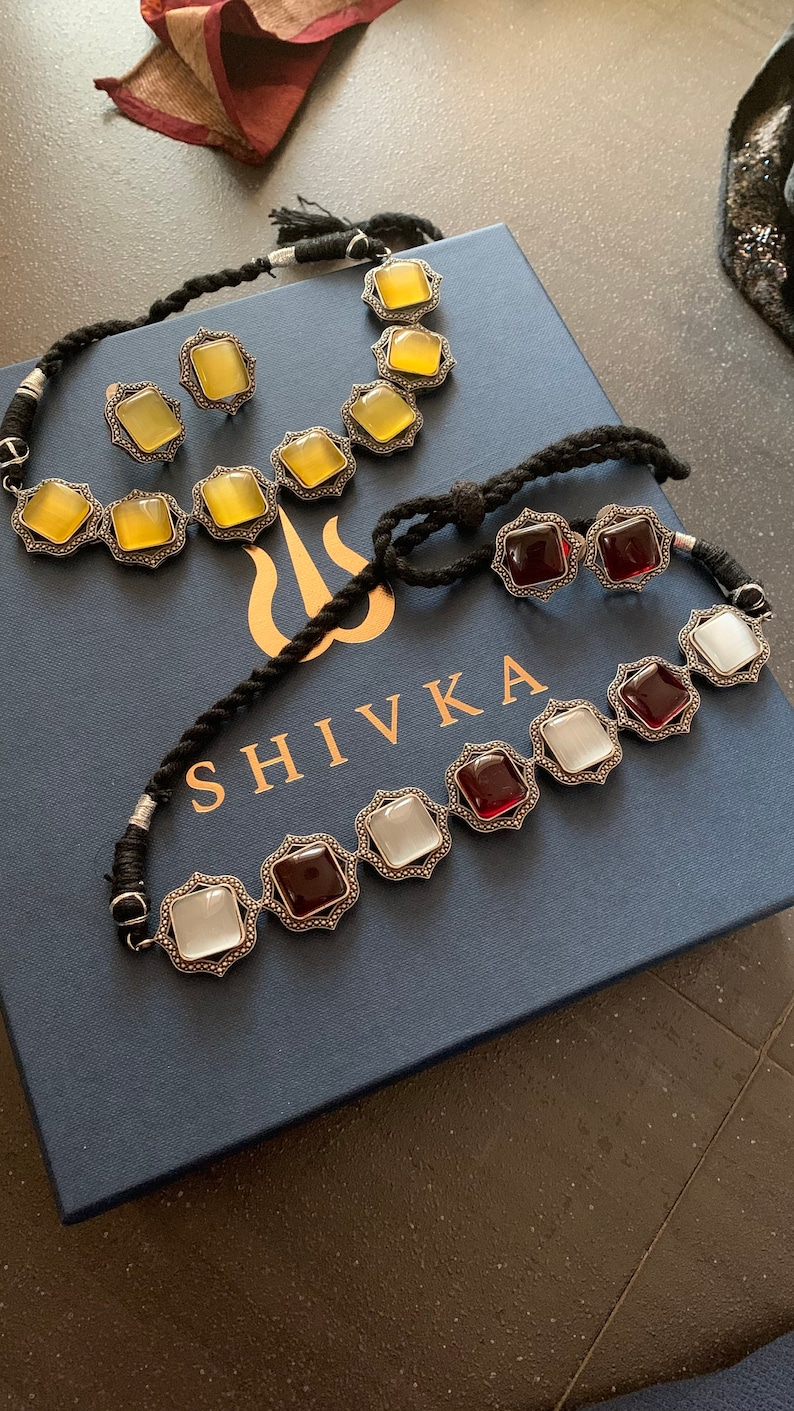 May include: Two silver necklaces with colorful gemstone accents. The first necklace has yellow gemstones and the second necklace has red and white gemstones. Both necklaces are on a blue box with the word "SHIVKA" printed on it.