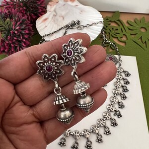 May include: Silver-toned jewelry set featuring a necklace and earrings. The earrings have a floral design with a small red stone in the center. The necklace has a delicate chain with floral and small bell-shaped charms.