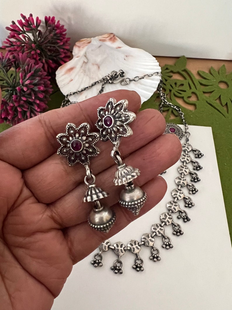 May include: Silver-toned jewelry set featuring a necklace and earrings. The earrings have a floral design with a small red stone in the center. The necklace has a delicate chain with floral and small bell-shaped charms.