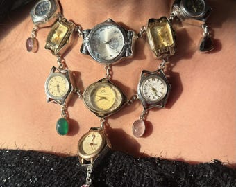 Könnte beinhalten: Eine einzigartige Halskette mit einer Sammlung von Vintage-Uhren in verschiedenen Formen und Größen. Die silberfarbene Kette hält die Uhren, einige mit goldfarbenen Zifferblättern, und ist mit bunten Edelsteinanhängern verziert. Ein auffälliges Schmuckstück.
