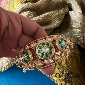 K&ouml;nnte beinhalten: Ein goldenes Armreif-Armband mit gr&uuml;nen und wei&szlig;en Steinen. Das Armband ist mit aufwendigen floralen Mustern verziert.