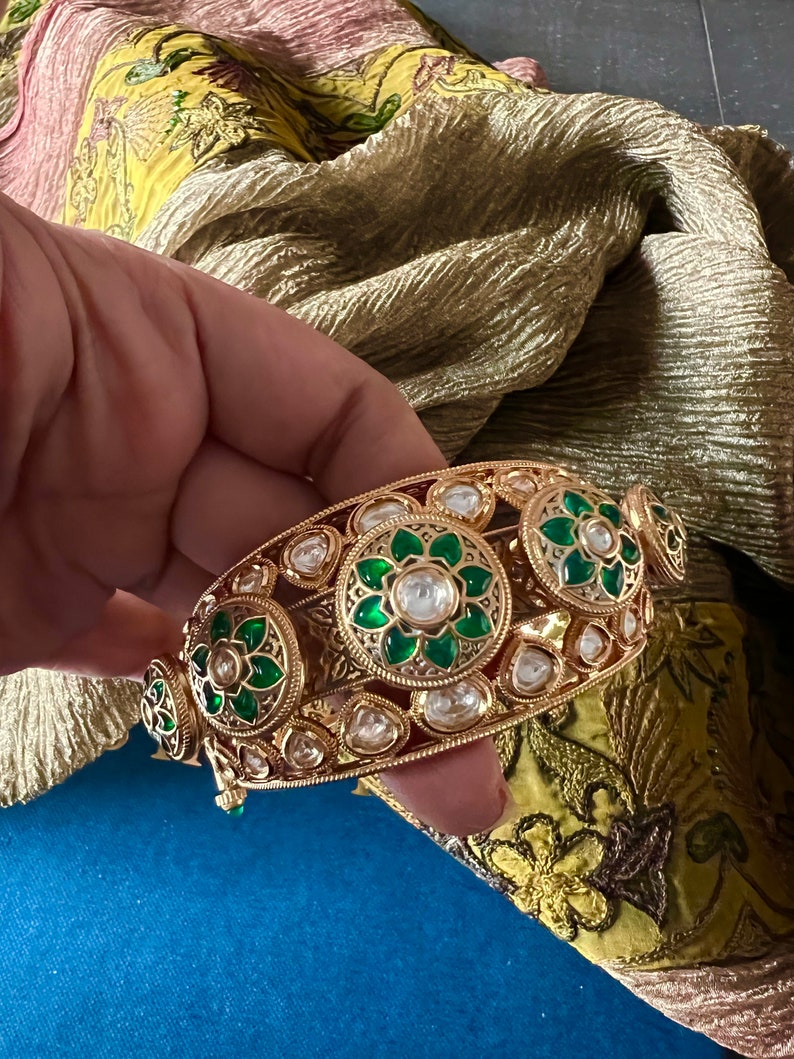 K&ouml;nnte beinhalten: Ein goldenes Armreif-Armband mit gr&uuml;nen und wei&szlig;en Steinen. Das Armband ist mit aufwendigen floralen Mustern verziert.