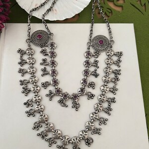 May include: A silver-toned necklace with a layered design. The necklace features intricate floral and geometric patterns, with small, round, red gemstones. The necklace is displayed on a white surface.