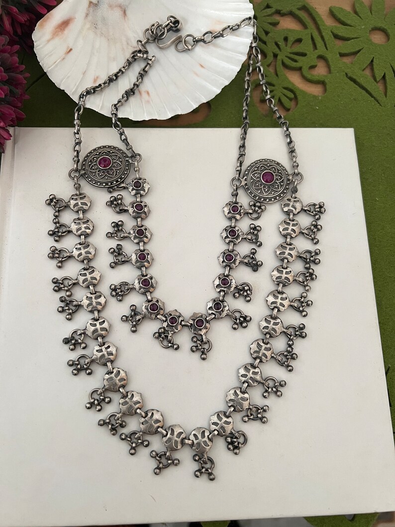 May include: A silver-toned necklace with a layered design. The necklace features intricate floral and geometric patterns, with small, round, red gemstones. The necklace is displayed on a white surface.