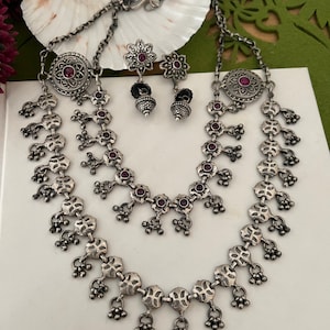 May include: A silver-toned necklace and earring set. The necklace features a layered design with small, round, red gemstones and dangling accents. The earrings have a floral design with a red gemstone center. The jewelry is displayed on a white surface.