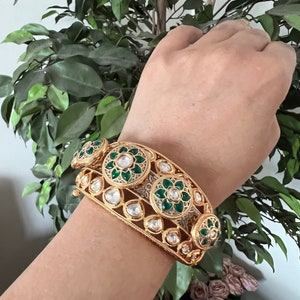 K&ouml;nnte beinhalten: Ein goldenes Armreif-Armband mit gr&uuml;nen und wei&szlig;en Emaille-Blumenmustern. Das Armband ist mit klaren Steinen verziert.