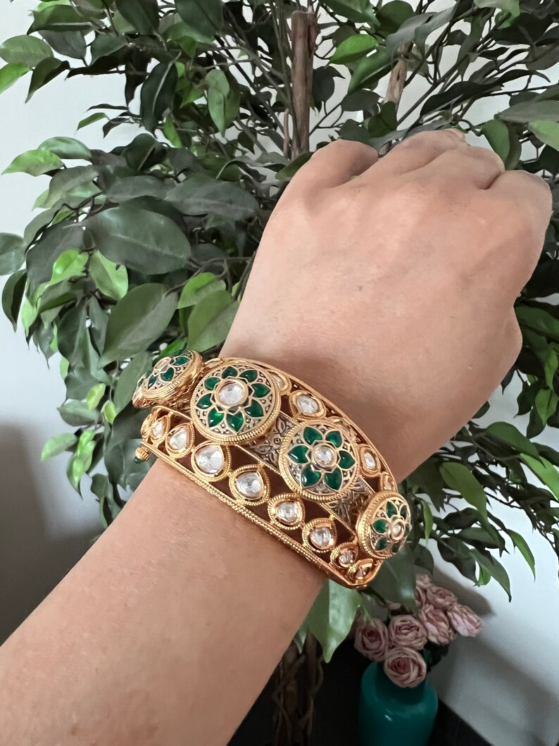 K&ouml;nnte beinhalten: Ein goldenes Armreif-Armband mit gr&uuml;nen und wei&szlig;en Emaille-Blumenmustern. Das Armband ist mit klaren Steinen verziert.