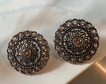 May include: A pair of silver earrings with a circular design. The earrings are adorned with small, clear rhinestones and have a delicate, intricate pattern.