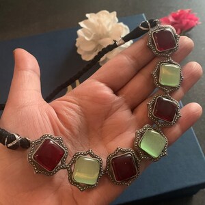 May include: A silver necklace with alternating red and green square-shaped stones. The necklace is made of a black cord.