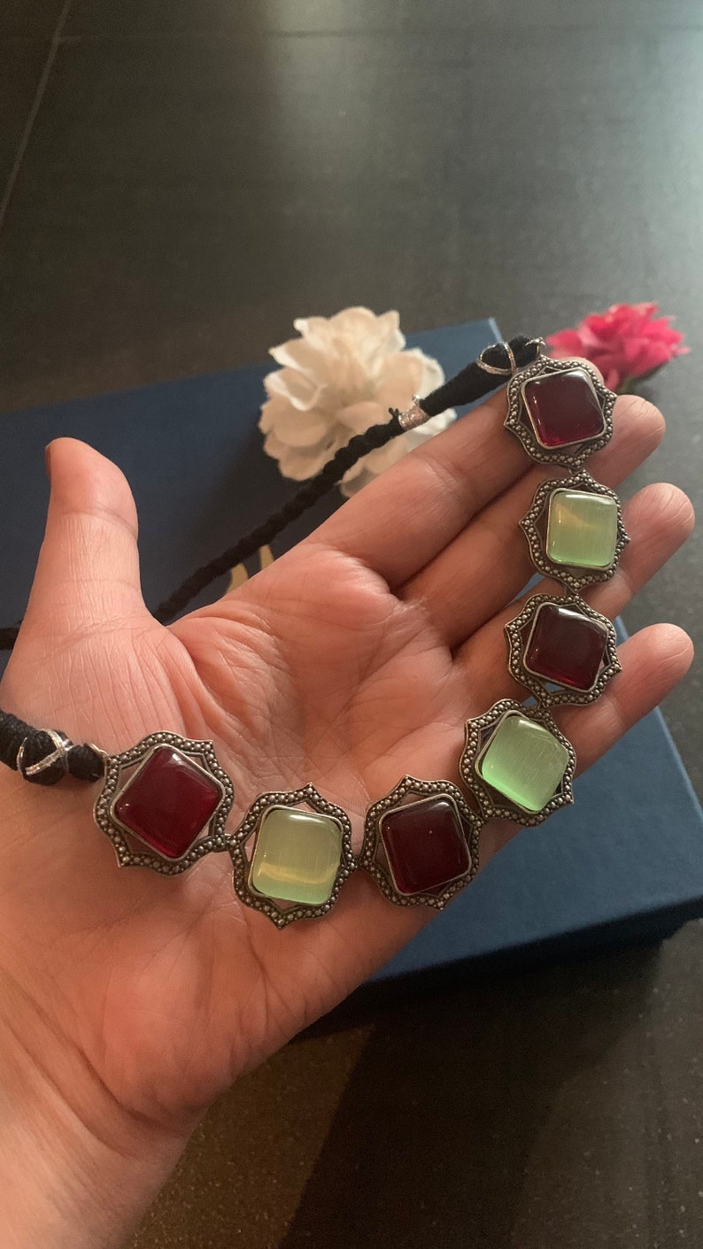 May include: A silver necklace with alternating red and green square-shaped stones. The necklace is made of a black cord.
