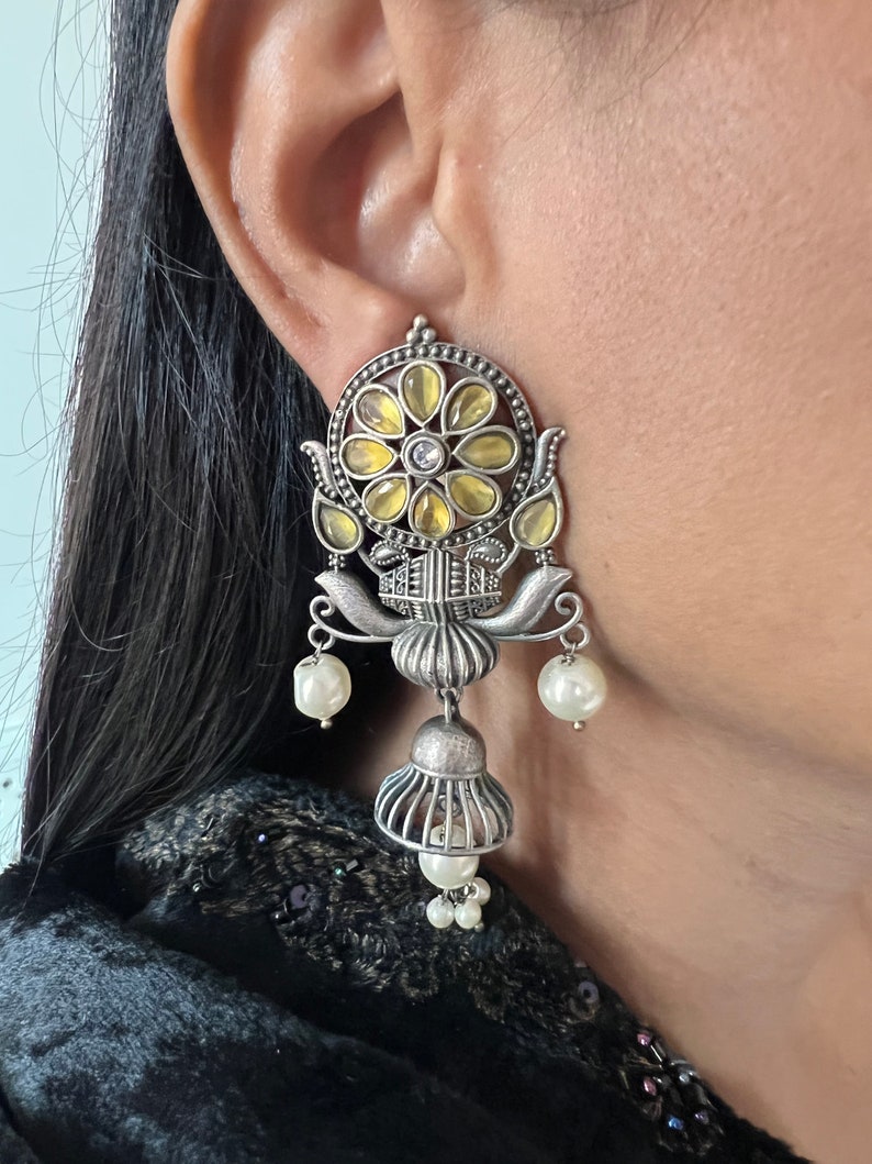 May include: Silver earrings with a floral design and yellow and white stones. The earrings have a bell-shaped dangle with pearl accents.