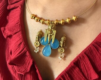 May include: A gold-toned necklace with a pendant featuring three light blue teardrop-shaped stones. The necklace has a beaded section and small dangling earrings with pearls. The background is a red top.