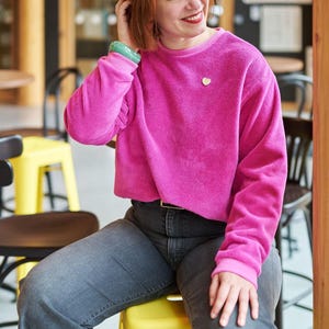 Puede incluir: Una persona con una sudadera corta rosa brillante con un broche de corazón dorado. También lleva vaqueros grises y una pulsera verde. El fondo incluye sillas amarillas y negras.