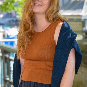 Könnte beinhalten: Eine Frau mit langen, welligen, rotbraunen Haaren trägt ein braunes ärmelloses Oberteil und einen dunkelblauen Cardigan. Sie trägt auch einen dunkelblauen Rock mit Knöpfen.