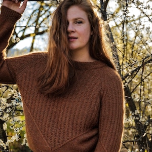 Könnte beinhalten: Eine Frau mit langen, roten Haaren trägt einen braunen Strickpullover. Sie steht vor einem Baum mit weißen Blüten.