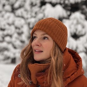 May include: A person wearing a rust-colored knit beanie and matching neck warmer. The person is also wearing a rust-colored puffer jacket. The background is a snowy forest.