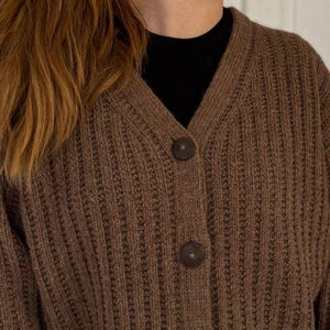 Könnte beinhalten: Ein brauner Strickcardigan mit V-Ausschnitt und Knopfleiste. Der Cardigan hat eine gerippte Struktur und ist aus einem weichen, warmen Material gefertigt.
