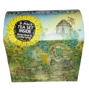 May include: A decorative box for a children's tea set, featuring a whimsical garden scene with a girl at a table. The box includes a 9-piece tea set, a recipe book, and a cookie cutter.