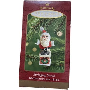 Può includere: Ornamento natalizio Hallmark Keepsake con una figura di Babbo Natale su una molla. L'ornamento è in una scatola con il testo "Springing Santa" e "DÉCORATION DES FÊTES". La figura di Babbo Natale ha barba bianca, abito rosso e base camino.