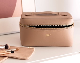 May include: A beige makeup case with a gold zipper and handle, personalized with the initials "HC". Two small beige pouches and makeup brushes are next to the case. A framed abstract art print is in the background.