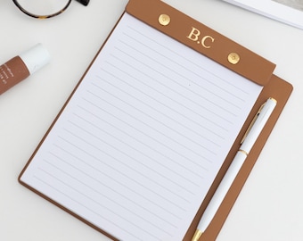 May include: A brown leather notepad with the initials "B.C." in gold lettering, a white pen, and a pair of tortoiseshell glasses rest on a white surface. A tube of "Summer Fri" is also present, along with an open magazine.