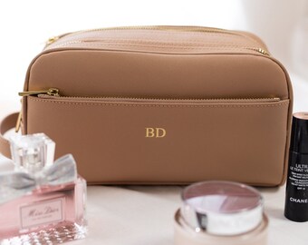May include: A tan cosmetic bag with gold zippers and the initials "BD" in gold. The bag is surrounded by makeup items, including a lipstick, perfume, a makeup brush, foundation, and powder.