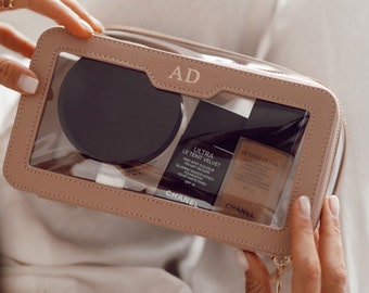May include: A light pink makeup bag with a clear front panel, containing a black compact and two bottles of Chanel makeup. The bag is personalized with the initials "AD" and has a gold zipper.