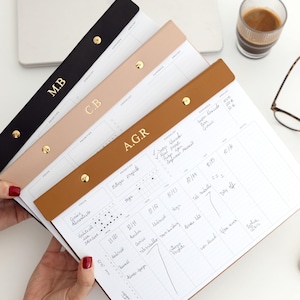 May include: A collection of personalised desk calendars with leather straps in black, beige, and brown, each with gold initials. The calendars are open to show the weekly planning pages. A laptop, coffee, and glasses are in the background.