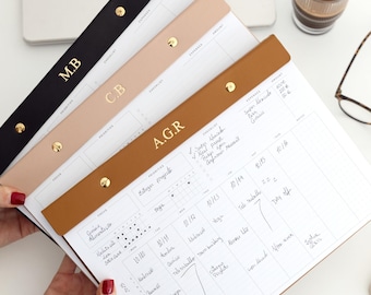 May include: A stack of personalized desk calendars with leather straps in black, beige, and brown, each with gold initials. The calendars are open to display the weekly planning pages. A laptop, coffee, and glasses are in the background.