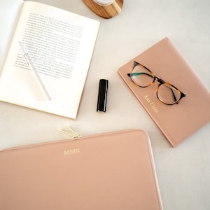 Pu&ograve; includere: Una custodia per laptop in pelle rosa chiaro con cerniera e le iniziali "MMR" in rilievo dorato. La custodia &egrave; su una superficie bianca accanto a un bicchiere d'acqua, un libro, un paio di occhiali e un rossetto.