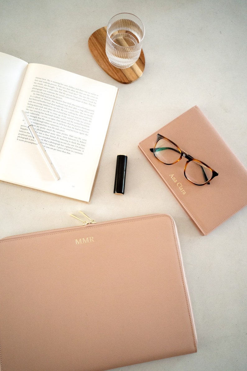Pu&ograve; includere: Una custodia per laptop in pelle rosa chiaro con cerniera e le iniziali "MMR" in rilievo dorato. La custodia &egrave; su una superficie bianca accanto a un bicchiere d'acqua, un libro, un paio di occhiali e un rossetto.