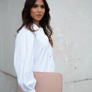 Pu&ograve; includere: Una donna con una camicia bianca abbottonata tiene una custodia per laptop in pelle rosa cipria con le iniziali "AT" in rilievo sul davanti.