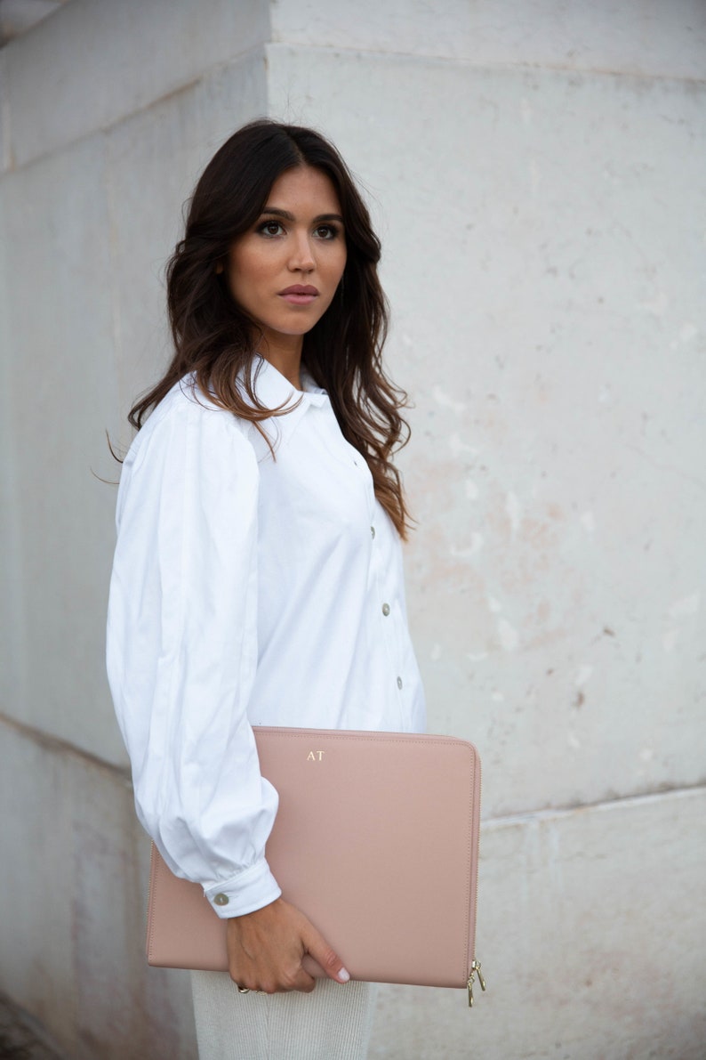 Pu&ograve; includere: Una donna con una camicia bianca abbottonata tiene una custodia per laptop in pelle rosa cipria con le iniziali "AT" in rilievo sul davanti.