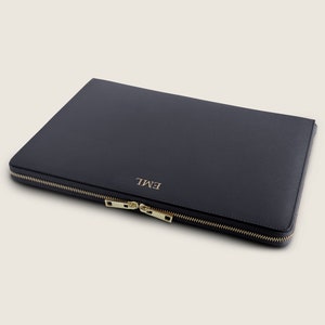 Pu&ograve; includere: Una custodia per laptop in pelle blu scuro con una cerniera dorata e il nome "EMIL" in rilievo sul davanti.