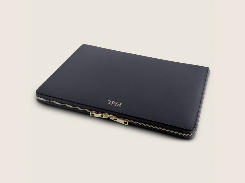 Pu&ograve; includere: Una custodia per laptop in pelle blu scuro con una cerniera dorata e il nome "EMIL" in rilievo sul davanti.