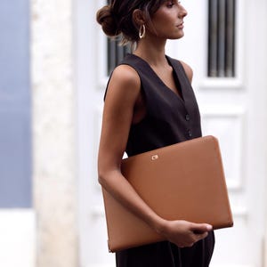 Pu&ograve; includere: Una donna con un vestito nero senza maniche tiene una custodia per laptop in pelle marrone con le iniziali "CB" in rilievo sul davanti.