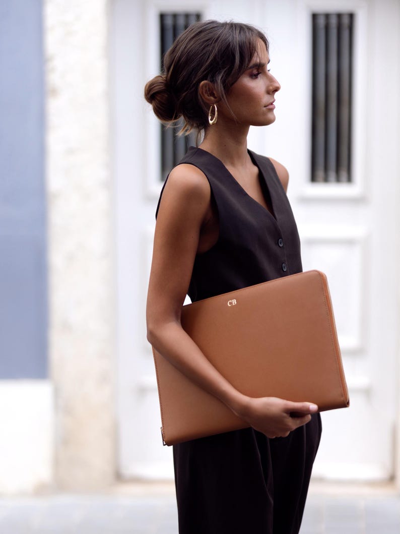 Pu&ograve; includere: Una donna con un vestito nero senza maniche tiene una custodia per laptop in pelle marrone con le iniziali "CB" in rilievo sul davanti.