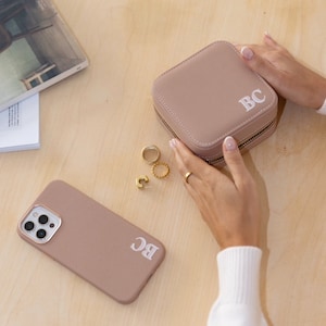 May include: A light pink leather jewellery box with a zipper closure and the initials "BC" embroidered on the top. The box is sitting on a wooden surface.