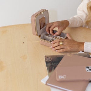 May include: A light pink jewellery box with a zip closure and a mirror inside. The box is open and filled with various rings and earrings. The box is sitting on a light wood table.