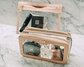 May include: Two clear plastic zippered makeup bags with pink trim. The smaller bag has the name "MIA" on it. The larger bag has the initials "CM" on it and contains various makeup and beauty products.