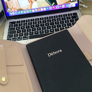 K&ouml;nnte beinhalten: Ein schwarzes Ledernotizbuch mit dem Namen "D&eacute;bora" in goldenen Lettern gepr&auml;gt. Das Notizbuch ist ge&ouml;ffnet und liegt auf einem hellrosa Lederorganizer mit mehreren Taschen und einem goldenen Knopf.