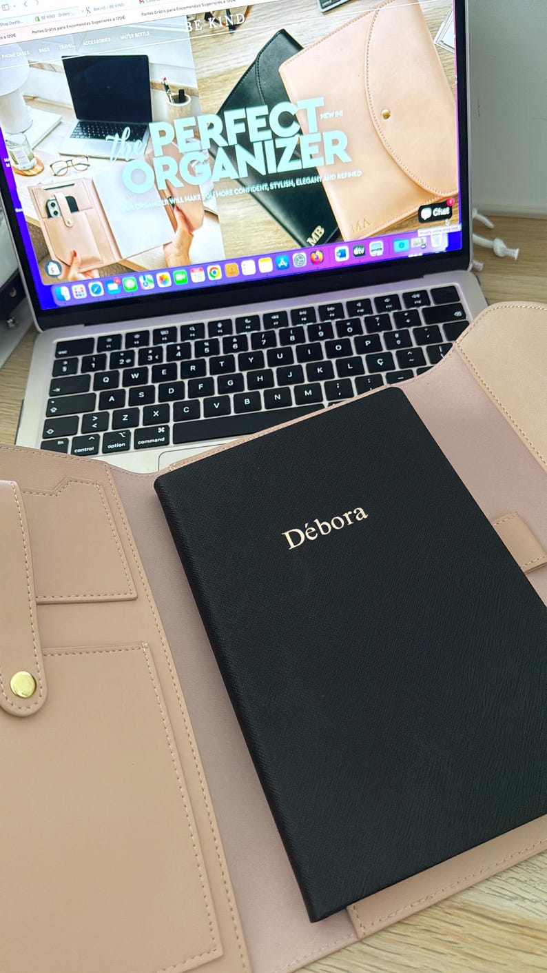 K&ouml;nnte beinhalten: Ein schwarzes Ledernotizbuch mit dem Namen "D&eacute;bora" in goldenen Lettern gepr&auml;gt. Das Notizbuch ist ge&ouml;ffnet und liegt auf einem hellrosa Lederorganizer mit mehreren Taschen und einem goldenen Knopf.