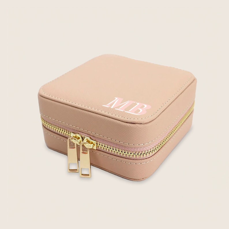 May include: A light pink square-shaped jewellery box with a gold zipper and the initials "MB" in white with a pink outline.