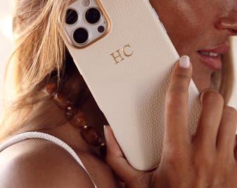 May include: Cream-colored phone case with gold-colored accents and the initials "HC" in gold. The case has a textured surface and is designed for a smartphone. The phone case is being held up to the ear.