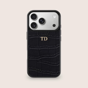 Puede incluir: Funda de teléfono negra con un patrón texturizado similar al de un cocodrilo. La funda tiene un marco negro y una sección de cámara plateada. Las letras doradas "TD" están centradas en la funda.