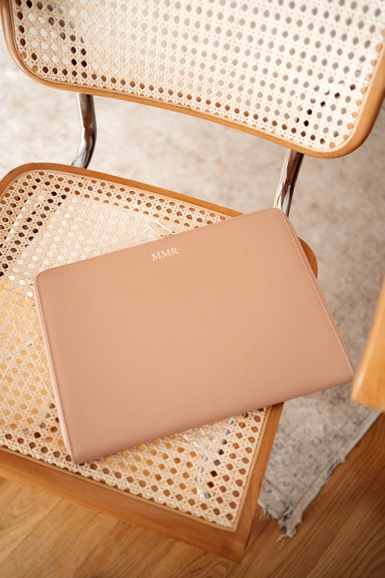 Pu&ograve; includere: Una custodia per laptop in pelle marrone chiaro con le iniziali "MMR" in rilievo in oro sul davanti. La custodia &egrave; appoggiata su una sedia intrecciata.